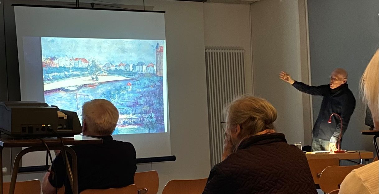 Der Kunsthistoriker Dr. Carstensen zeigt mit ausgestrecktem Arm auf ein an die Wand projeziertes Bild. Im Vordergrund sitzt ein interessiertes Publikum.