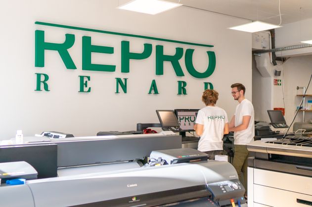 Das Logo von Renard in großen grünen Buchstaben an einer Wand. Im Vordergrund der Produktionsraum mit Großen Druckern.
