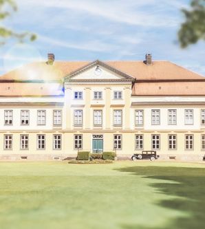 Historisches Herrenhaus mit symmetrischer Fassade und rotem Dach, umgeben von gepflegtem Rasen und Bäumen.