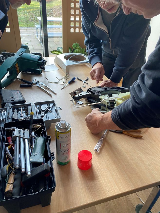 Auf dem Bild sieht man drei Personen, die sich über ein zerlegtes Küchengerät beugen, um dieses zu reparieren.