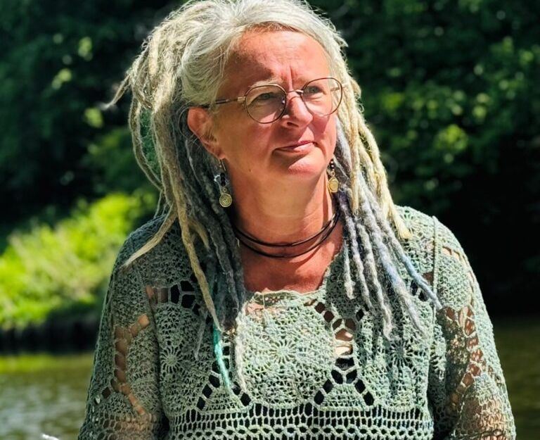 Ein Foto der Autorin Insa Ritterhoff im hellgrünen Oberteil vor einem See und grünen Bäumen im Hintergrund. Sie hält eine Kladde und einen Stift in der Hande. Ihre ergrauten Haare trägt sie lang in Dreads im Zopf.