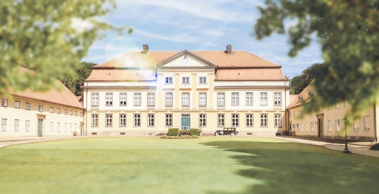 Historisches Herrenhaus mit symmetrischer Fassade und rotem Dach, umgeben von gepflegtem Rasen und Bäumen.