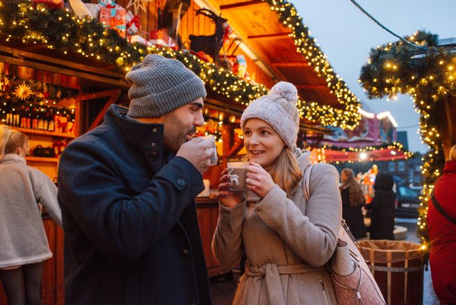 Ein Paar steht an einem festlich geschmückten Weihnachtsmarkt und hält Tassen in der Hand. Sie lächeln sich an und genießen die winterliche Atmosphäre.
