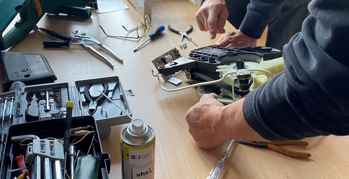 Auf dem Bild sieht man drei Personen, die sich über ein zerlegtes Küchengerät beugen, um dieses zu reparieren.