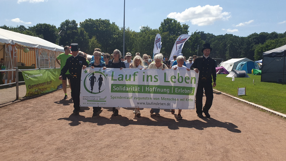 Mehrere Personen gehen gemeinsam über einen Weg im Freien und tragen ein Banner mit der Aufschrift „Lauf ins Leben“, im Hintergrund sind Zelte und Bäume zu sehen.