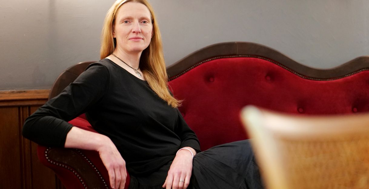 Die Erzählerin Frauke Freise sitzt seitlich  auf einem eleganten roten Sofa und schaut frontal in die Kamera. Sie trägt schwarz und die langen Haare offen. Die Hände liegen entspannt über der Lehne und über den Beinen.