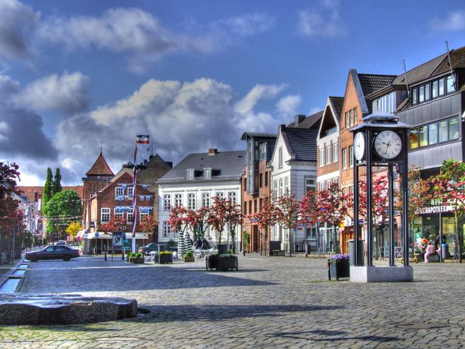 Ein Marktplatz mit historischen Gebäuden, einer großen Uhr und blühenden Bäumen unter einem blauen Himmel mit Wolken.