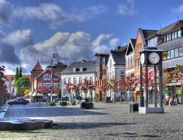 Ein Marktplatz mit historischen Gebäuden, einer großen Uhr und blühenden Bäumen unter einem blauen Himmel mit Wolken.